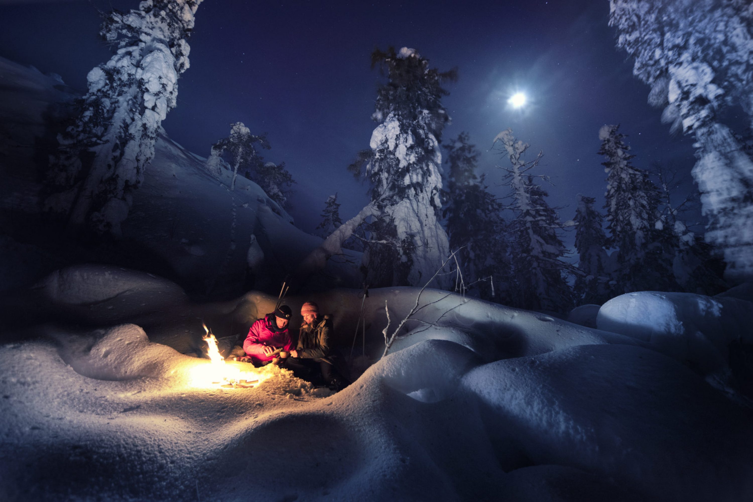 Pariskunta istuu lumessa nuotion ääressä yötaivaan alla