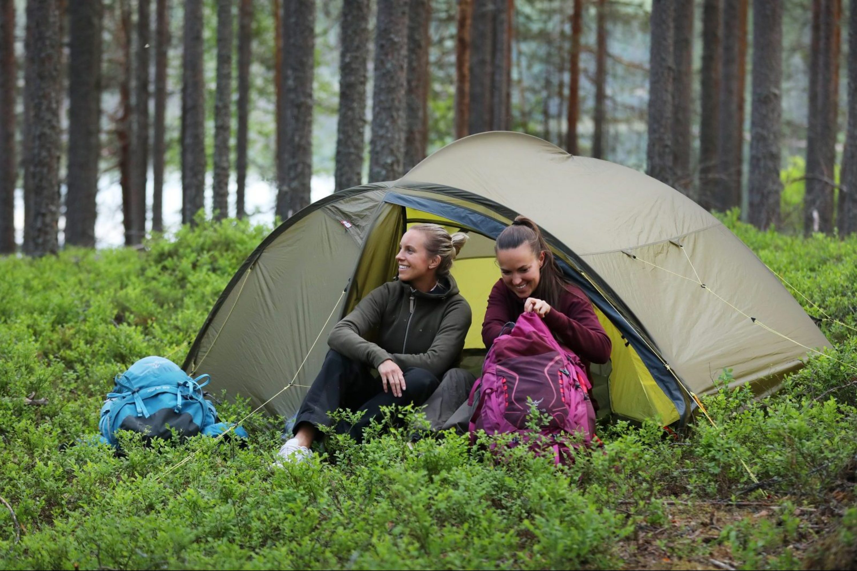 Kaksi naista istuvat teltan edessä metsässä
